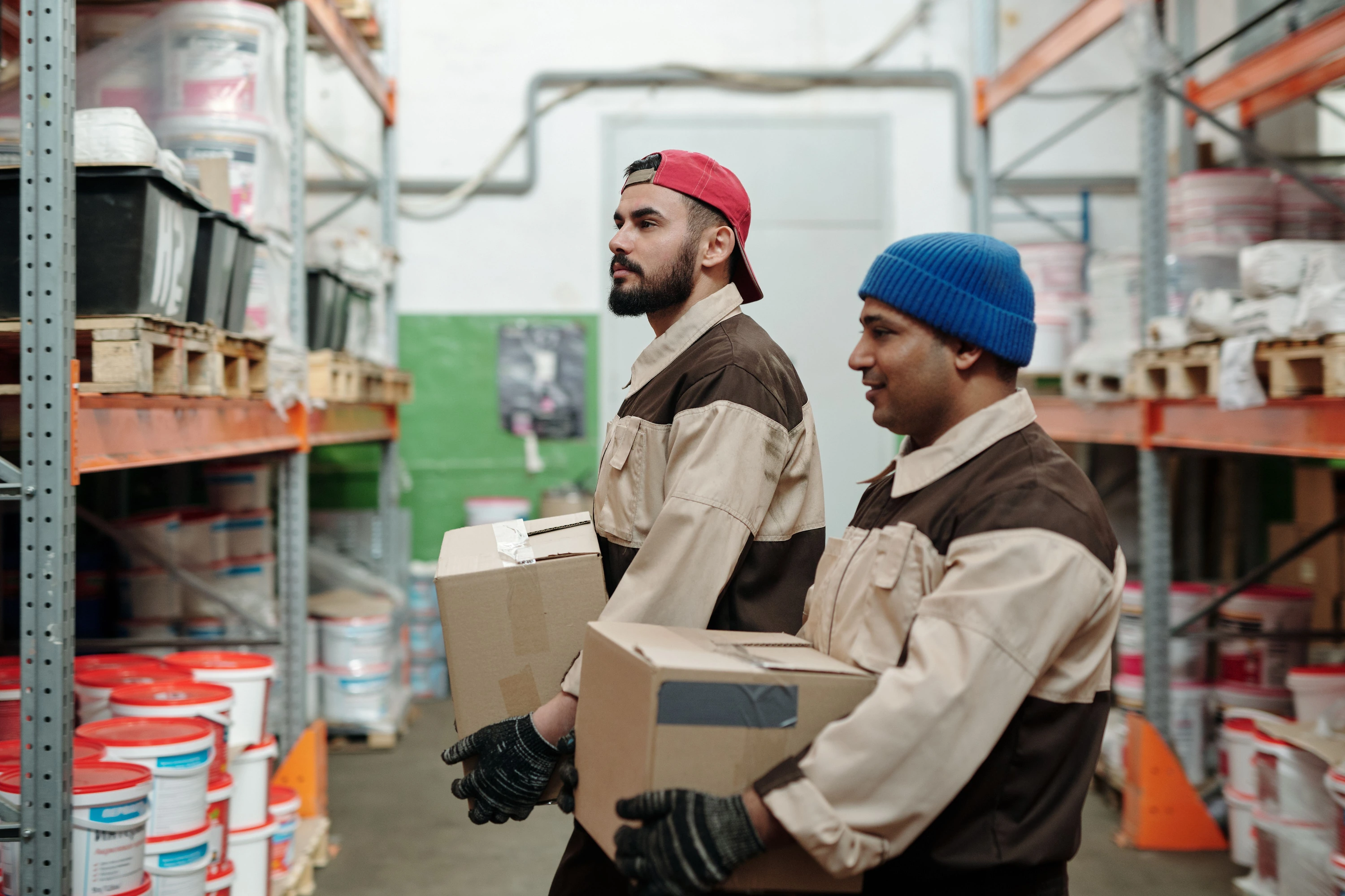 Two warehouse workers carrying boxes along stocked shelves